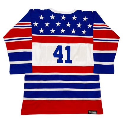 New York Americans hockey jersey. White collar. Blue shoulders with white stars, Alternating red, white, and blue bands of various sizes on sleeves and on bottom half of jersey. Large white band below with #41 in blue. Royal Retros